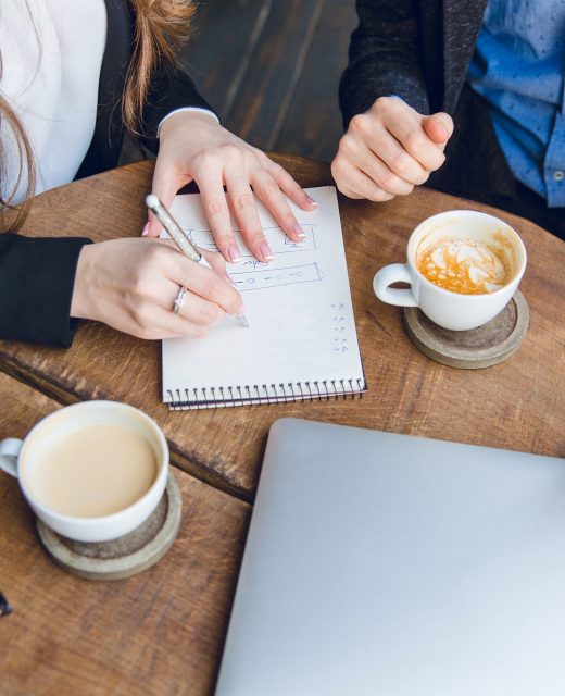 Close-up of a coffee table with two colleagues - man and woman - writing in notebook. Two cups of coffee, smartphone, laptop and eyeglasses are on the table. Woman wears silver ring and writes in notebook.